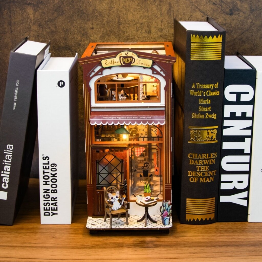 Miniatúrna drevená kaviarň s mačkou sediacou v kresle sa nachádza na drevenej poličke medzi knihami. Knihy obsahujú názvy: „Design Hotels Year Book09“, „A Treasury of World's Classics“, „Charles Darwin The Descent of Man“ a „Century“.