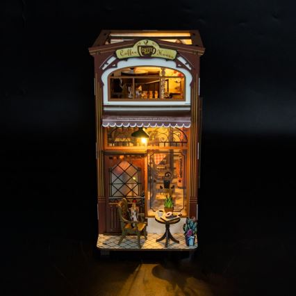 Drevená miniatúra kaviarne s osvetlením zobrazuje vnútorný priestor s nábytkom a stolom. Na fasáde je nápis „Coffee House“. „Measuring the happy pieces of life with a Coffee Saloon.“