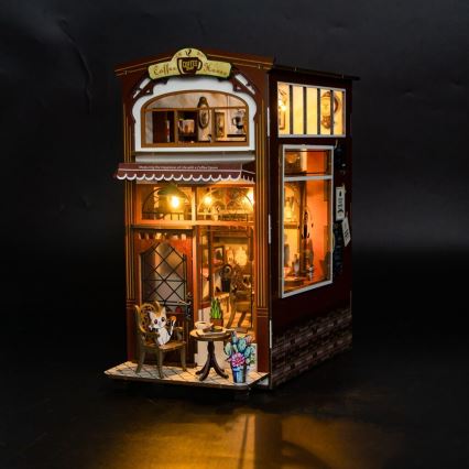 Drevená miniatúrna kaviarň s názvom „Coffee House“ osvetlená vnútri. Na terase sedí mačka pri stole. Kaviarňu zdobia kvety a drobné detaily.