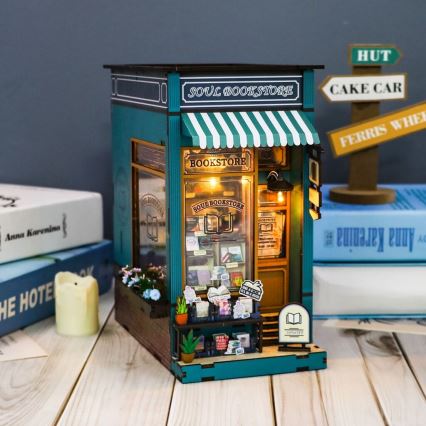 Miniatúrny model kníhkupectva „Soul Bookstore“ s výkladnou skriňou a regálmi s knihami, umiestnený na drevenom stole vedľa kníh a smajlíkových značiek.