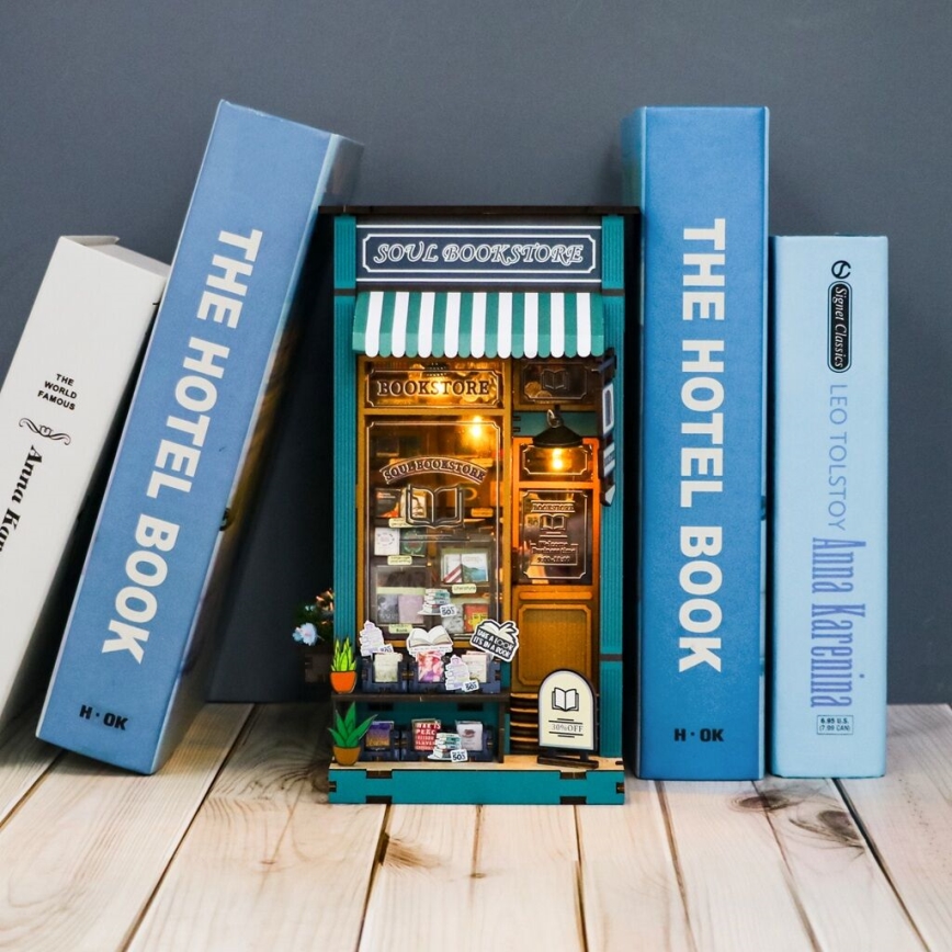 Miniatúrny kníhkupectvo „SOUL BOOKSTORE“ – stojacie medzi knihami „THE HOTEL BOOK“ a „Anna Karenina“ na drevenom povrchu.