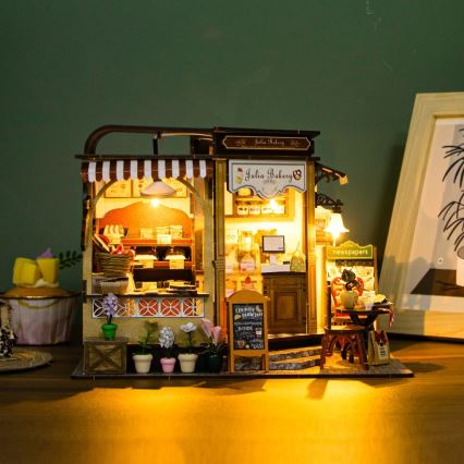 Drevená miniatúra pekárne „Julia Bakery“ s osvetlením, ponúkajúca pečivo a noviny, je umiestnená na drevenom stole. Na tabuli je text: „Cookies, Cupcakes, Brownie, Bread“.