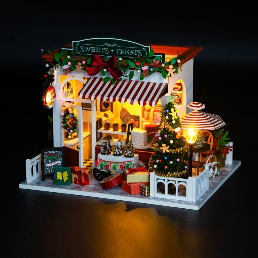 Miniatúrny cukráreň - osvetlený interiér zobrazuje sladkosti a vianočný stromček. Vonku sú darčeky. Nápis znie: „Sweets + Treats“.