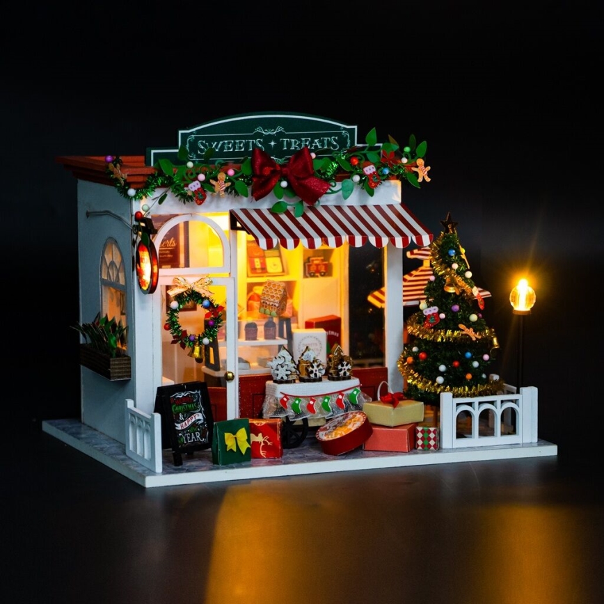 Miniatúrny vianočný obchodík s cukrovinkami a pochúťkami je osvetlený, pred ním je vianočný stromček a darčeky. Na tabuli je text: Christmas & Happy New Year.