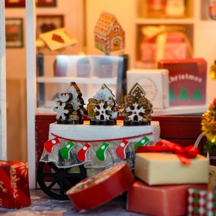 Vianočný vozík s perníkovými ozdobami v tvare stromčekov a snehových vločiek, zdobený vianočnými pančuchami, stojí v miniatúrnom vianočnom obchode obklopenom darčekovými krabičkami a vianočnými dekoráciami.