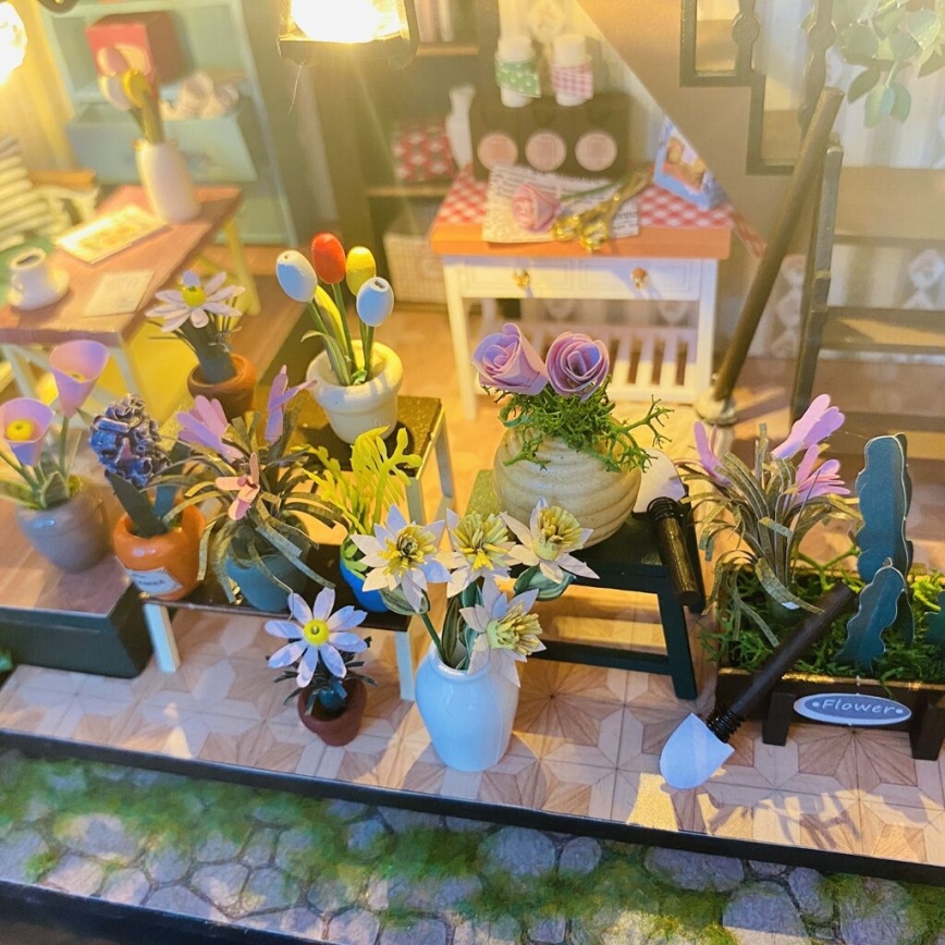 Miniatúrne kvety v kvetináčoch a vázach sú usporiadané na drevených stolíkoch v malej kvetinárstve. Na jednom drevenom boxe je nápis „Flower“. V pozadí je viditeľná časť interiéru s nábytkom.