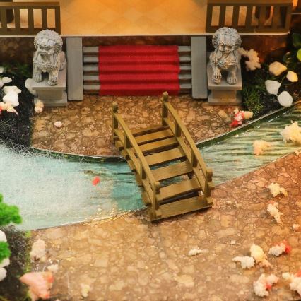 Drevený mostík prechádza cez miniatúrnu vodnú plochu. Mostík sa nachádza v miniatúrnej záhradnej scéne s kamennými sochami a červeným kobercom na schodoch.