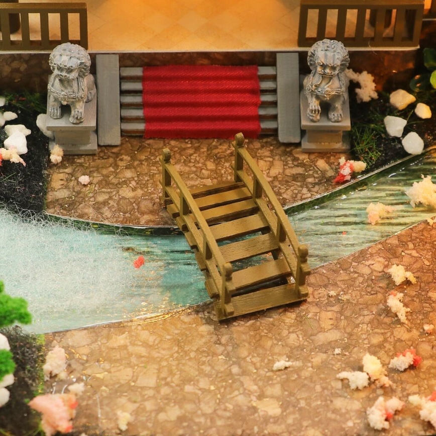 Drevený mostík prechádza cez miniatúrnu vodnú plochu. Mostík sa nachádza v miniatúrnej záhradnej scéne s kamennými sochami a červeným kobercom na schodoch.