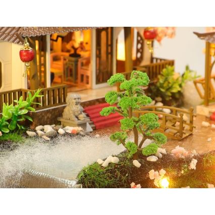 Miniatúrny stromček sa nachádza v záhrade miniatúrneho domu s čínskym štýlom. V pozadí je vidieť domček s osvetleným interiérom a mostíkom.