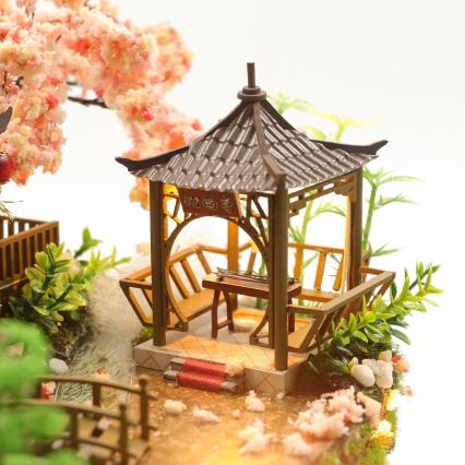 Miniatúrny altánok s hudobným nástrojom stojí v miniatúrnej záhrade s kvetmi čerešne a mostíkom. Na altánku je nápis.