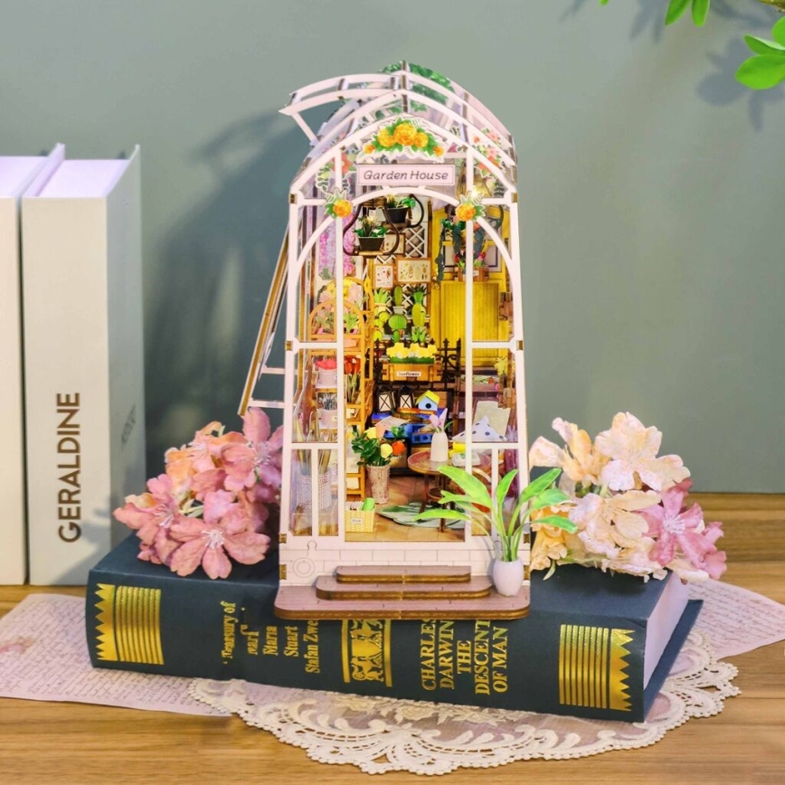 Miniatúrny skleníkový domček, stojaci na knihách, je obklopený umelými kvetmi. Na skleníku je nápis „Garden House“.