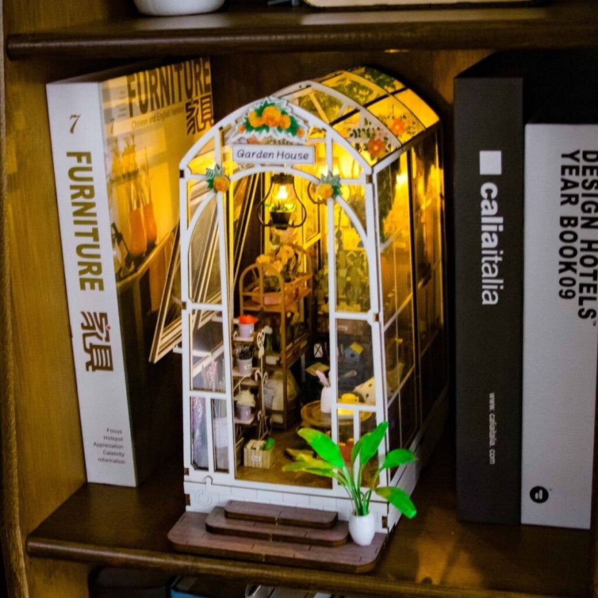 Miniatúrny sklenený skleník s vnútorným zariadením a osvetlením stojí na drevenej poličke medzi knihami „Furniture 家具“ a „Design Hotels. Year Book 09“. Na schodíkoch pred skleníkom je malá rastlina v bielom kvetináči.