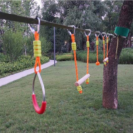 Lanová dráha s rôznymi prekážkami, ako sú drevené tyče a kruhy, je zavesená medzi stromami v záhrade. Prekážky sú pripevnené k hlavnému lanu pomocou šnúrok a karabín.