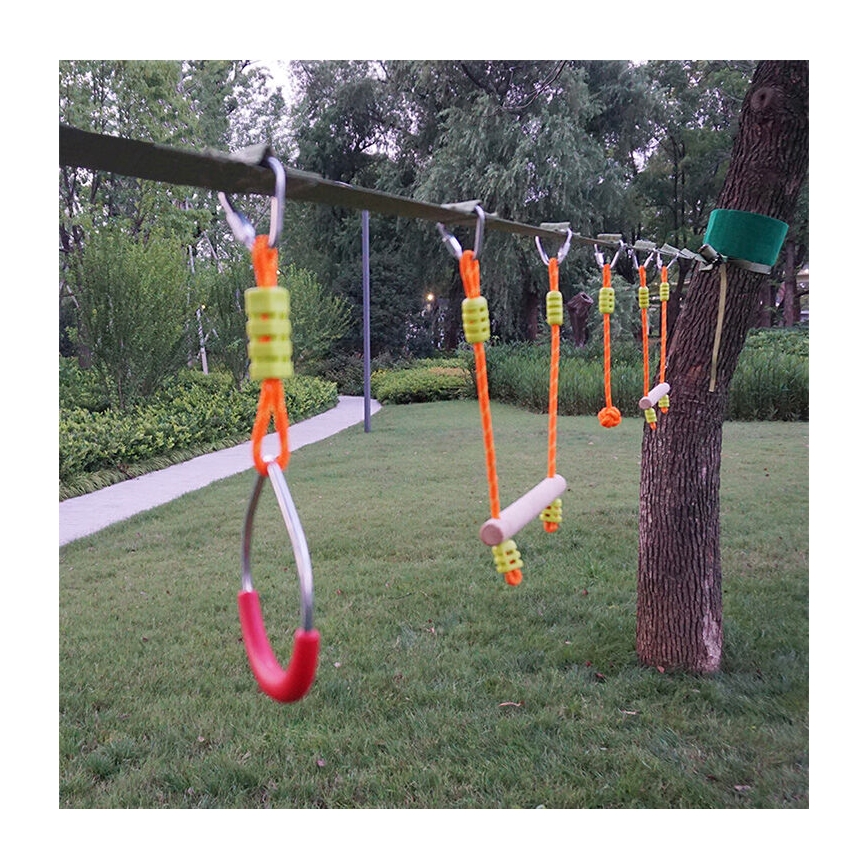 Lanová dráha s rôznymi prekážkami, ako sú drevené tyče a kruhy, je zavesená medzi stromami v záhrade. Prekážky sú pripevnené k hlavnému lanu pomocou šnúrok a karabín.