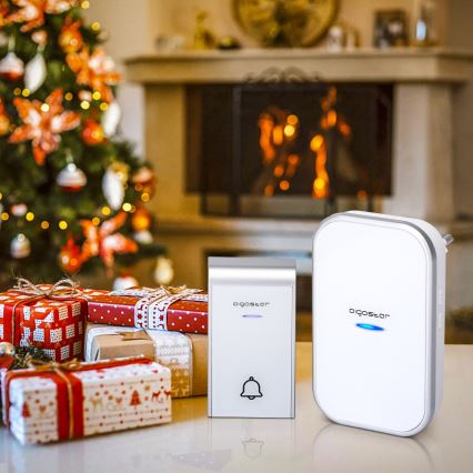 Dve bezdrôtové zvončekové jednotky Digostor, jedna väčšia a jedna menšia, sú umiestnené na stole pred rozmazaným pozadím vianočného stromčeka a krbu.