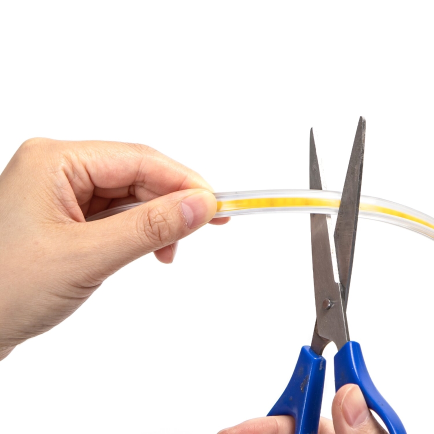 Ruka drží pružný svetelný zdroj s vnútorným žltým pruhom. Nožnice s modrými rukoväťami strihajú tento zdroj. pozadie je biele.