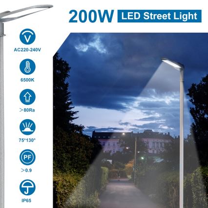 200W LED pouličné svetlo svieti v nočnom prostredí. Svetlo je osadené na stĺpe a osvetľuje chodník vedúci popri stromoch a budovách. Parametre: AC220-240V, 6500K, 80Ra, 75*130°, 0.9 PF, IP65.