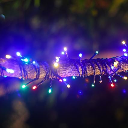 Elektrické svetielka rôznych farieb sú omotané okolo konára stromu v tme. Svietia modrou, červenou, zelenou a žltou farbou. Konár je hnedý a nachádza sa v exteriéri.