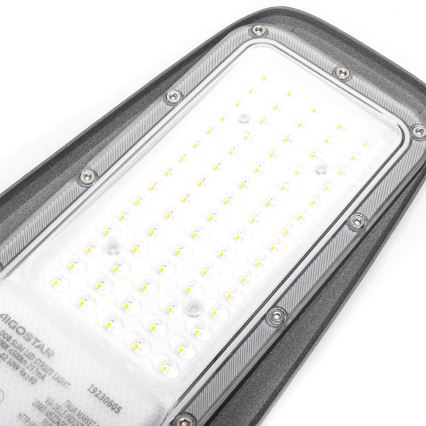 LED pouličné osvetlenie svieti. Je umiestnené na bielom pozadí. Na tele je nápis: AIGOSTAR DOB SLIM LED STREET LIGHT 50W Ra≥80 19230605 VIA DELLE INDUST 20883 MEZZAGO ITALIA MARKET.