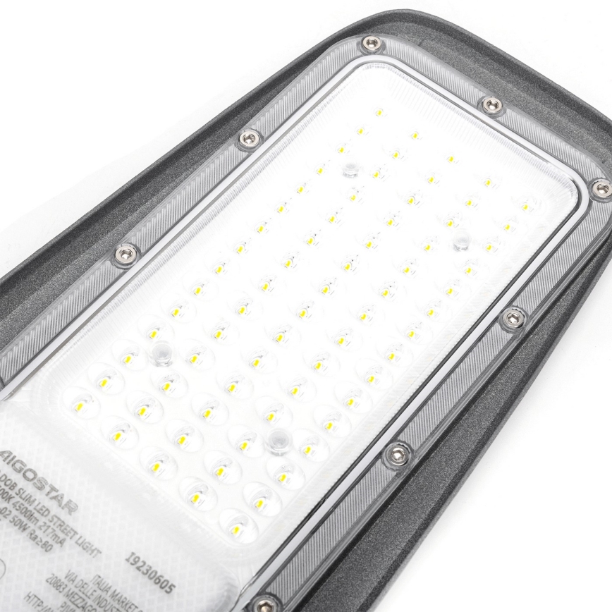 LED pouličné osvetlenie svieti. Je umiestnené na bielom pozadí. Na tele je nápis: AIGOSTAR DOB SLIM LED STREET LIGHT 50W Ra≥80 19230605 VIA DELLE INDUST 20883 MEZZAGO ITALIA MARKET.