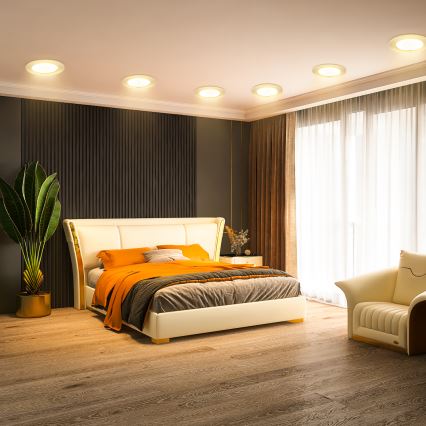 Posteľ s oranžovou prikrývkou stojí v spálni. V pozadí sú viditeľné stropné svetlá a závesy. Vpravo je umiestnené kreslo.