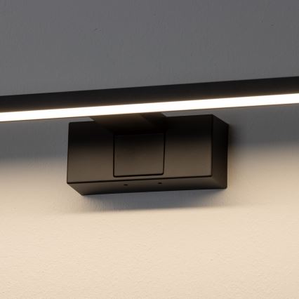 Čierna LED nástenná lampa svieti na stene. Lampa je pripevnená k stene pomocou čierneho hranolového držiaka. Svetlo je dlhé a úzke.