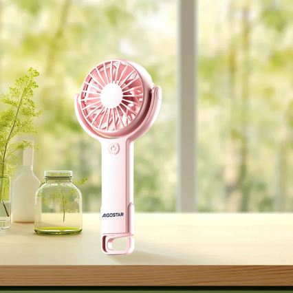 Ručný ventilátor značky AIGOSTAR stojí na drevenom stole pred oknom. Ventilátor je ružovej farby a je v nepoužívanom stave. Na pozadí je rozmazané zelené pozadie s oknom.