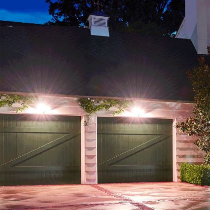 Dvojité zelené garážové vráta sú osvetlené dvoma vonkajšími svetlami. Vráta sú umiestnené na budove s tmavozelenou strechou a svetlými stenami. Okolité prostredie tvorí nočná scéna s vegetáciou.