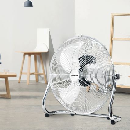 Stolový ventilátor Agostar stojí v svetlej miestnosti s drevenou stoličkou a nábytkom. Ventilátor je chromovaný s bielymi lopatkami.