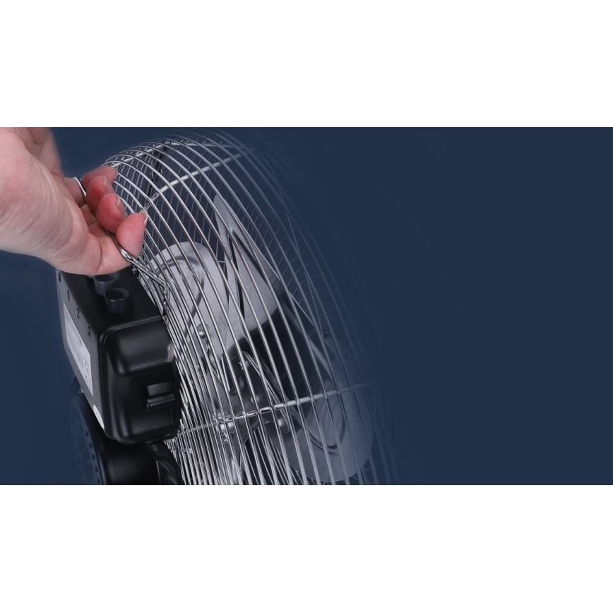 Ruka drží kovový ochranný kryt stolného ventilátora. Ventilátor má kovové lopatky a čierny plastový základ. Pozadie je tmavomodré.