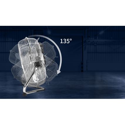 Kovový stolový ventilátor s kovovou ochrannou mriežkou sa otáča o 135°. Stojí na podlahe v tmavom, priemyselnom prostredí s veľkou rolovacou bránou v pozadí.
