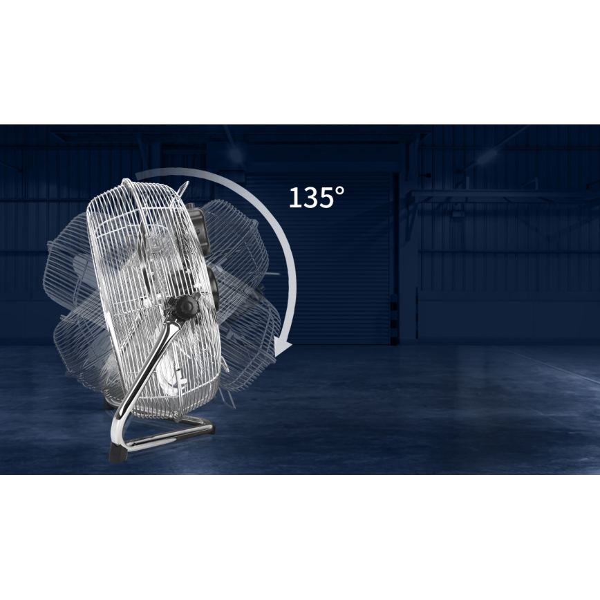 Kovový stolový ventilátor s kovovou ochrannou mriežkou sa otáča o 135°. Stojí na podlahe v tmavom, priemyselnom prostredí s veľkou rolovacou bránou v pozadí.