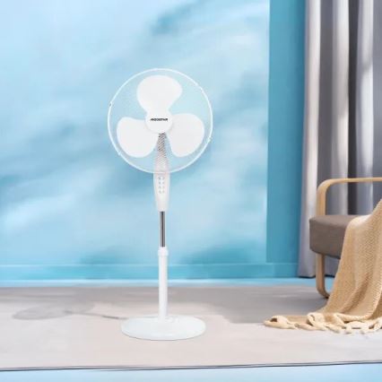 Biely stolový ventilátor značky AEGostar stojí na podlahe v svetlom interiéri. Ventilátor je zapnutý. V pozadí je vidieť svetlomodrú stenu, časť koberca a kreslo.