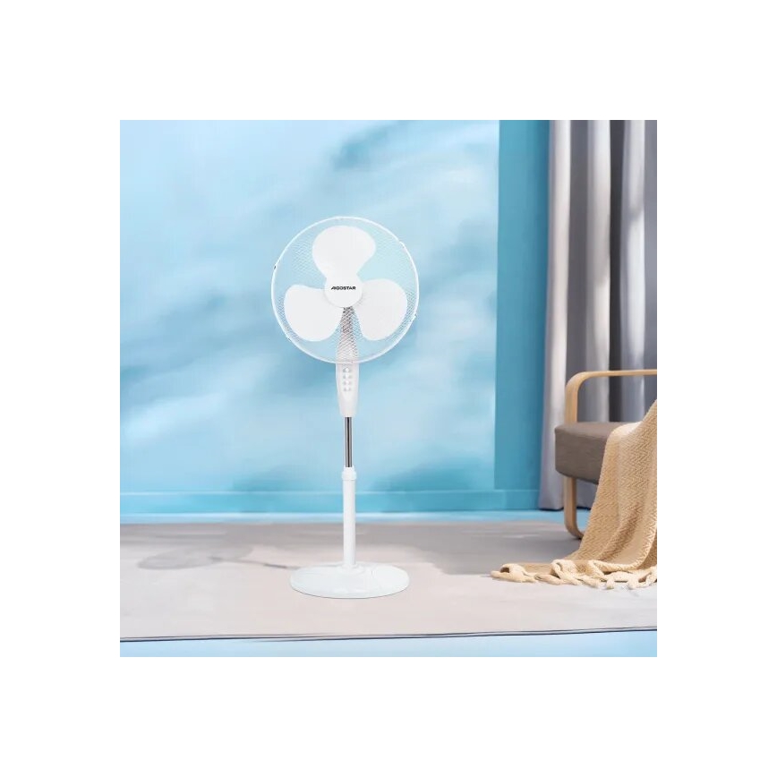 Biely stolový ventilátor značky AEGostar stojí na podlahe v svetlom interiéri. Ventilátor je zapnutý. V pozadí je vidieť svetlomodrú stenu, časť koberca a kreslo.