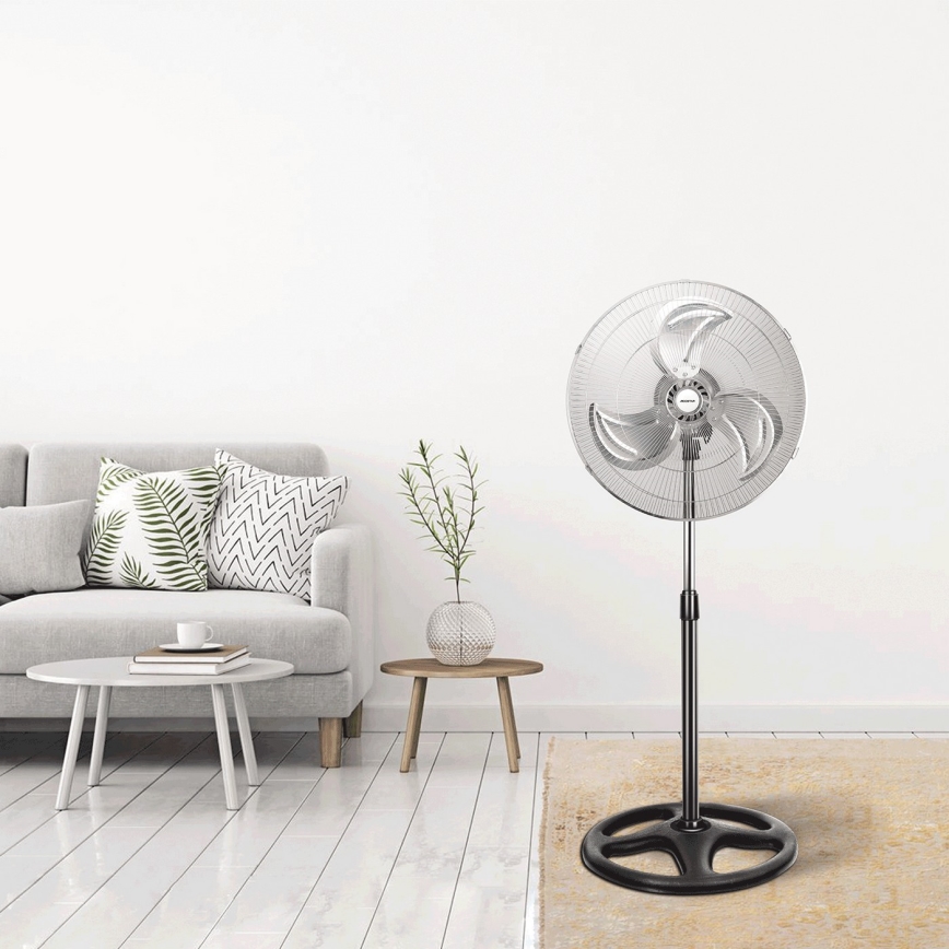 Stolový ventilátor stojí v obývacej izbe. Ventilátor je zapnutý. Nachádza sa na svetlej podlahe pred šedou sedačkou a nízkym stolíkom.