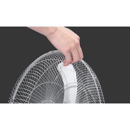 Ruka drží kovový ochranný kryt stojanového ventilátora pred bielymi vrtuľami. Ventilátor je na tmavšom pozadí.