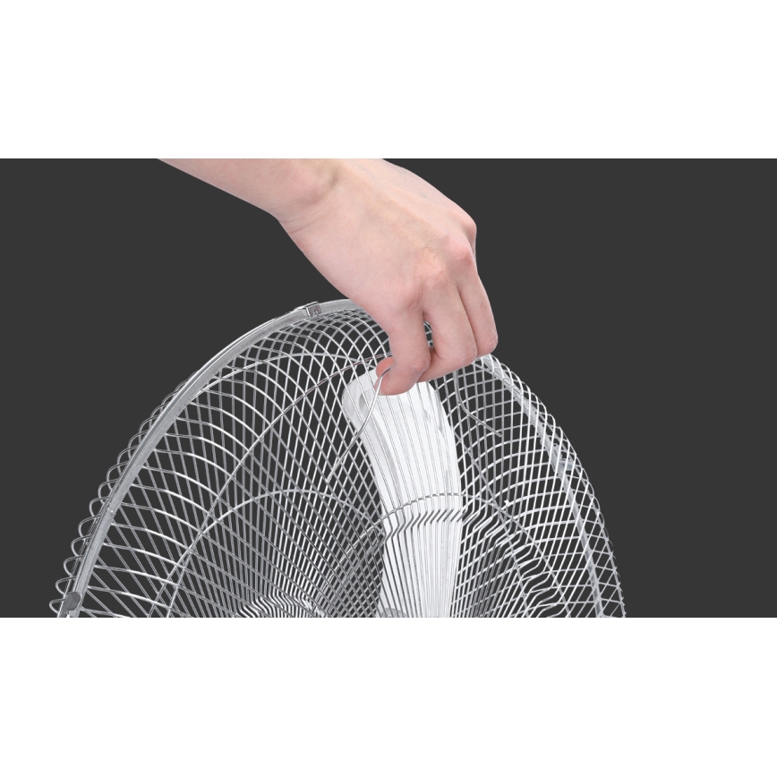 Ruka drží kovový ochranný kryt stojanového ventilátora pred bielymi vrtuľami. Ventilátor je na tmavšom pozadí.