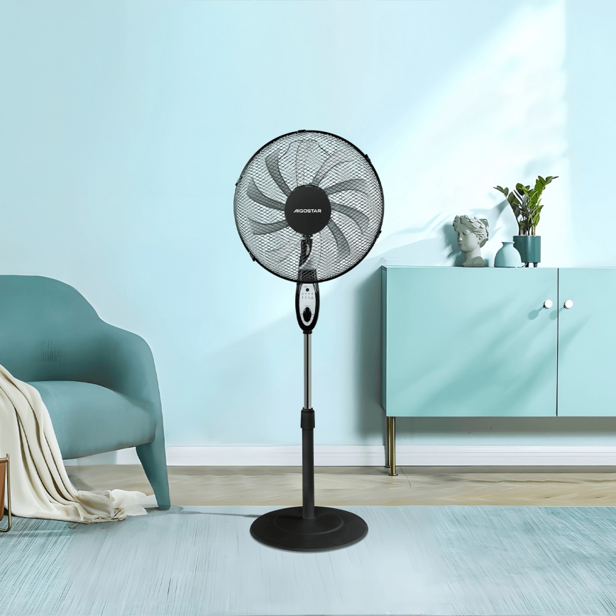Stolový ventilátor s kovovou mriežkou a nastaviteľnou výškou stojí v obývacej izbe. Ventilátor je umiestnený na podlahe pred sedačkou a komodou.
