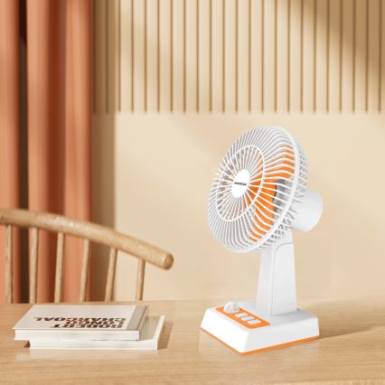Malý stolový ventilátor s oranžovými detailmi stojí na drevenom stole. Ventilátor je zapnutý. V pozadí je vidieť drevenú stoličku a záves.