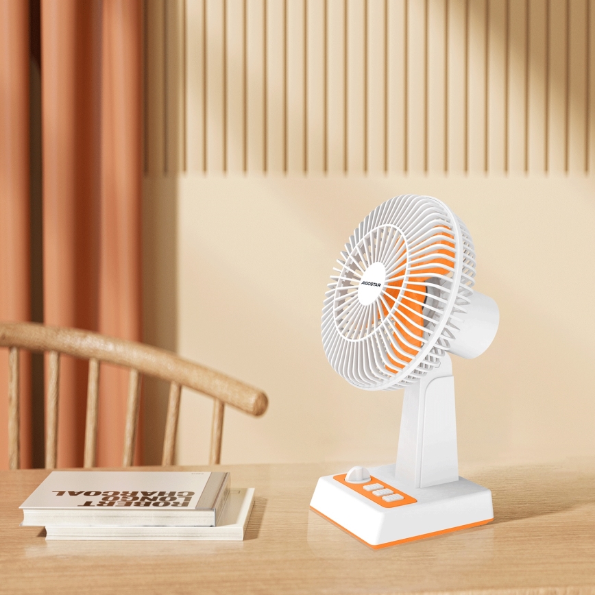 Malý stolový ventilátor s oranžovými detailmi stojí na drevenom stole. Ventilátor je zapnutý. V pozadí je vidieť drevenú stoličku a záves.