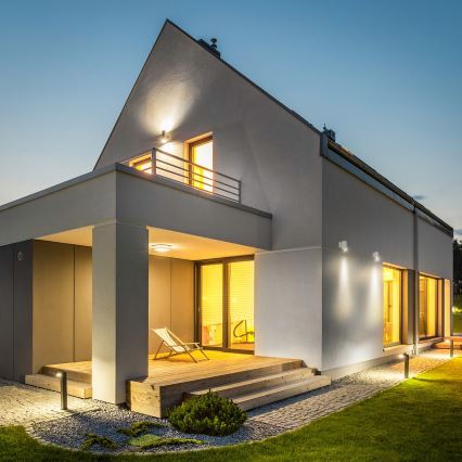 Moderná vila, večer osvetlená, má drevenú terasu s ležadlom. Vonkajšie osvetlenie zvýrazňuje jej architektúru.