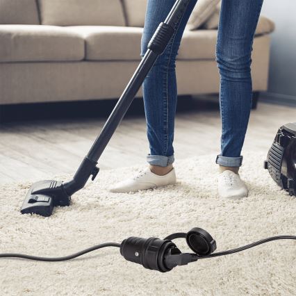 Žena vysáva koberec vysávačom. Na podlahe leží elektrický kábel s vidlicou. Okolím je obývacia izba s gaučom a drevenou podlahou.