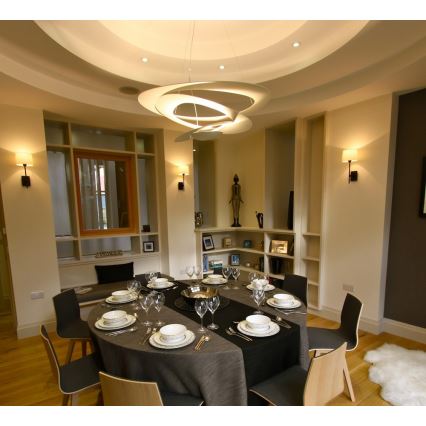 Jedálenský stôl s prestieraním je pripravený na jedlo v miestnosti s vstavanými knižnicami a moderným osvetlením. Svetlá stropná lampa dominuje priestoru.