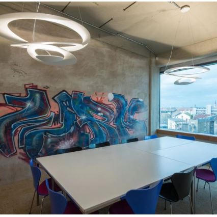 Dve moderné kruhové svietidlá osvetľujú konferenčný stôl v miestnosti s graffiti na stene a výhľadom na mesto cez veľké okno.