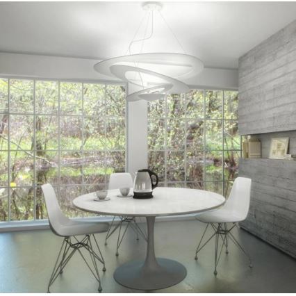 Moderná závesná lampa s abstraktnými krivkami visí nad bielym okrúhlym jedálenským stolom. Pri stole stoja štyri biele stoličky. Kontext tvorí svetlá miestnosť s veľkými oknami a betónovou stenou.