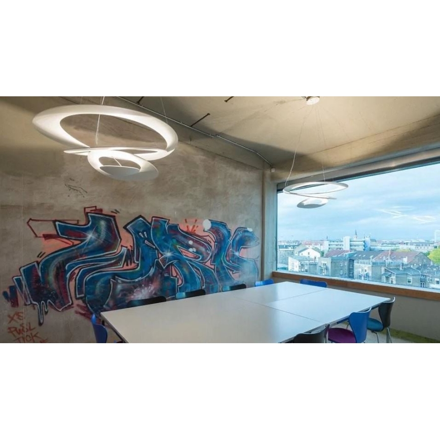 Dve moderné biele závesné lampy svietia v miestnosti s veľkým oknom. Stôl s kreslami sa nachádza pri stene s nástennou maľbou. Vidno mestskú panorámu.