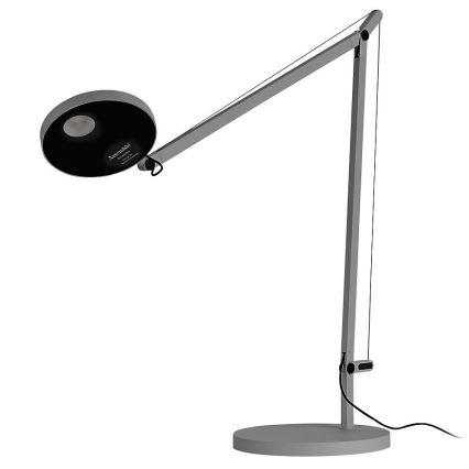 Stolová lampa s nastaviteľným ramenom a okrúhlou tienidlom svieti. Je sivá, na jednoduchej podstave, s viditeľným káblom. Na tienidle je nápis: Artemide, Design by Richard Sapper, Made in Italy.