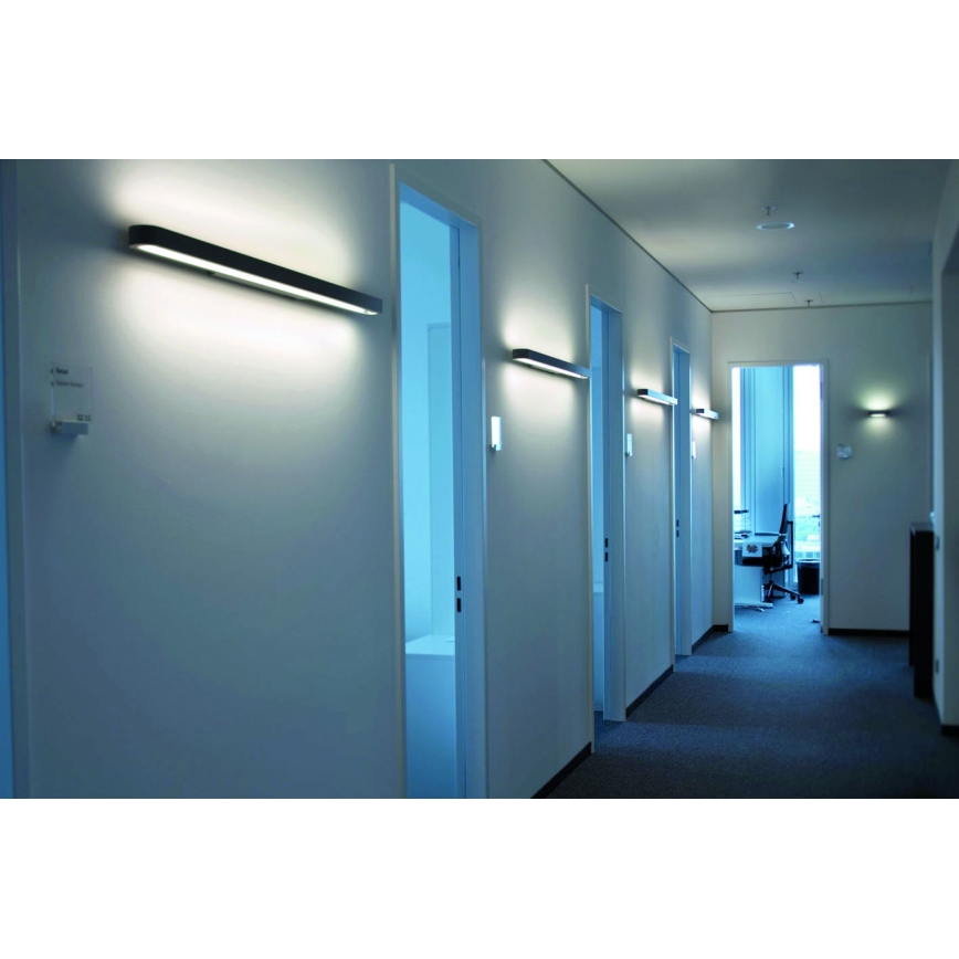 Svetlá svietia na stene dlhej chodby kancelárie. Dlhá obdĺžniková svietidlá sú pripevnené na stenu oproti dverám kancelárií. Chodba vedie k otvorenému priestoru.