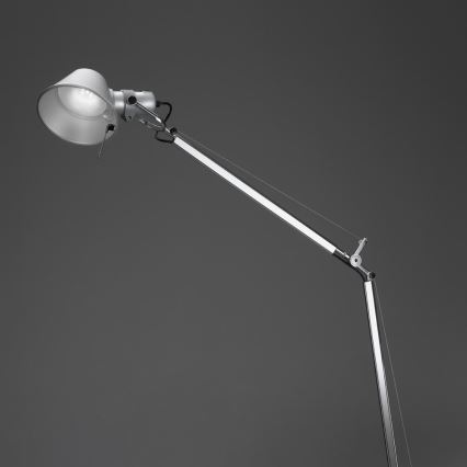 Stolová lampa s kovovou konštrukciou a nastaviteľným ramenom svieti v tmavošedej miestnosti.