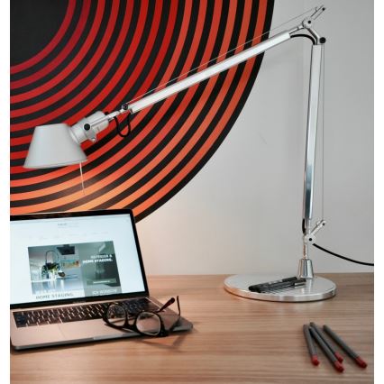 Strieborná stolová lampa s nastaviteľným ramenom svieti na drevenom stole. Na stole je notebook s otvorenou webovou stránkou s textom „WASSER HOME STAGING, REDESIGN & HOME STAGING, ICH WÜNSCHE“. Na notebooku ležia okuliare. Na stole sú aj pero a ceruzky. V pozadí je stena s červeno-čiernym kruhovým vzorom.